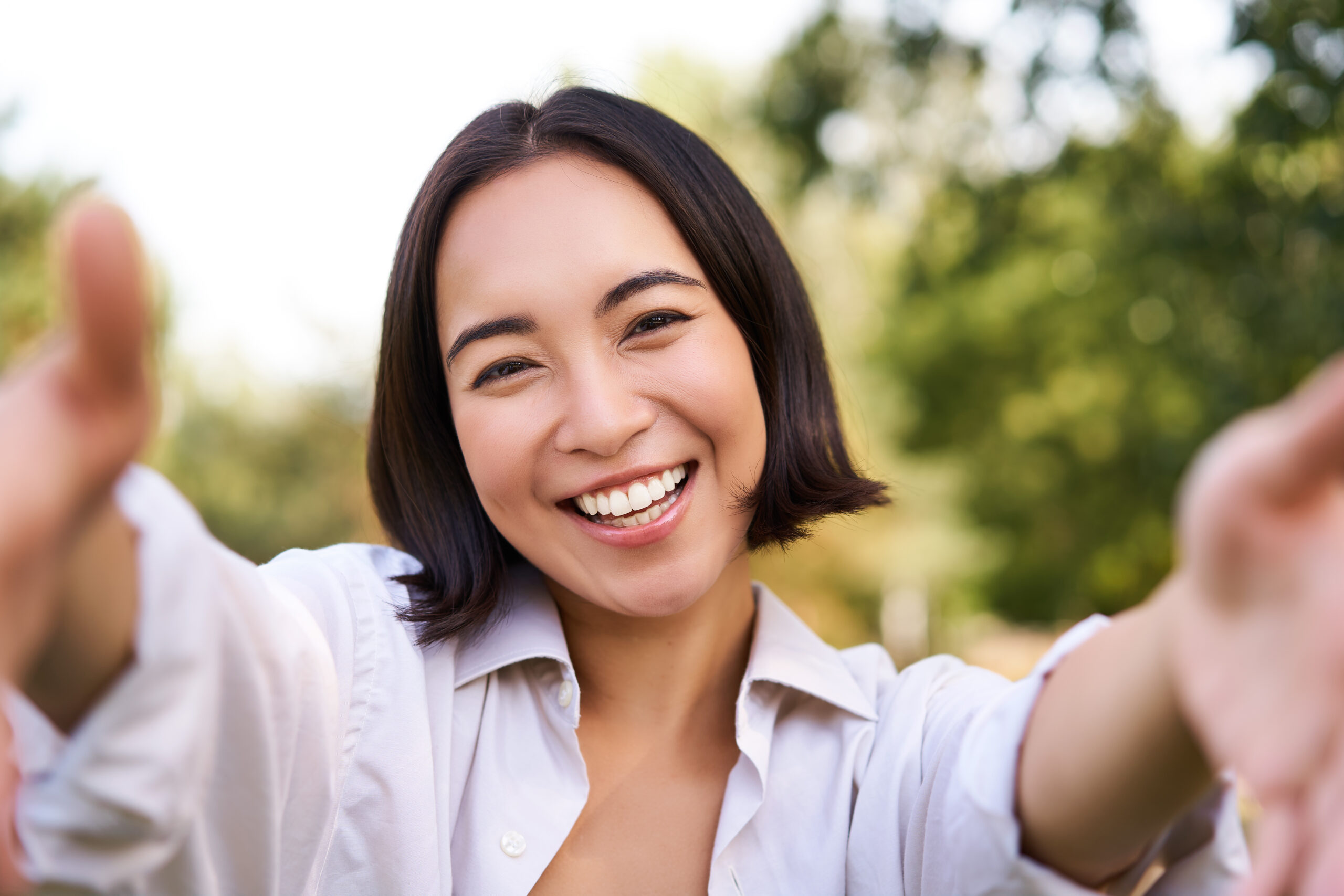 Smiling asian girl takes selfie, video chats, holds smartphone and speaks at camera, posing in park